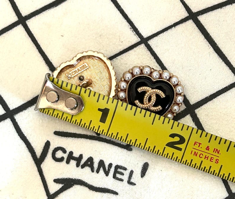 Pode incluir: Dois brincos Chanel em forma de cora&ccedil;&atilde;o. Um &eacute; dourado com o logotipo Chanel e o outro &eacute; preto com um logotipo dourado e rodeado por p&eacute;rolas falsas. Os brincos medem aproximadamente 2,5 cm de comprimento.