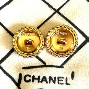 May include: Two round gold-tone metal buttons featuring a twisted rope border. Each button displays the text CHANEL at the top and the interlocking CC logo at the bottom. A central shank is visible on the back of each button, which is positioned over a white fabric surface with black grid lines and the text CHANEL.
