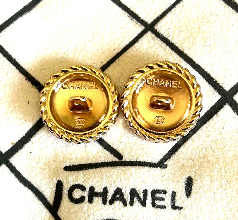 May include: Two round gold-tone metal buttons featuring a twisted rope border. Each button displays the text CHANEL at the top and the interlocking CC logo at the bottom. A central shank is visible on the back of each button, which is positioned over a white fabric surface with black grid lines and the text CHANEL.