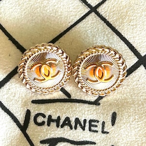 May include: A pair of round gold-tone earrings featuring a central interlocking CC logo surrounded by a sunburst pattern and a braided rope border. The earrings are displayed against a textured off-white fabric with the black printed text CHANEL.