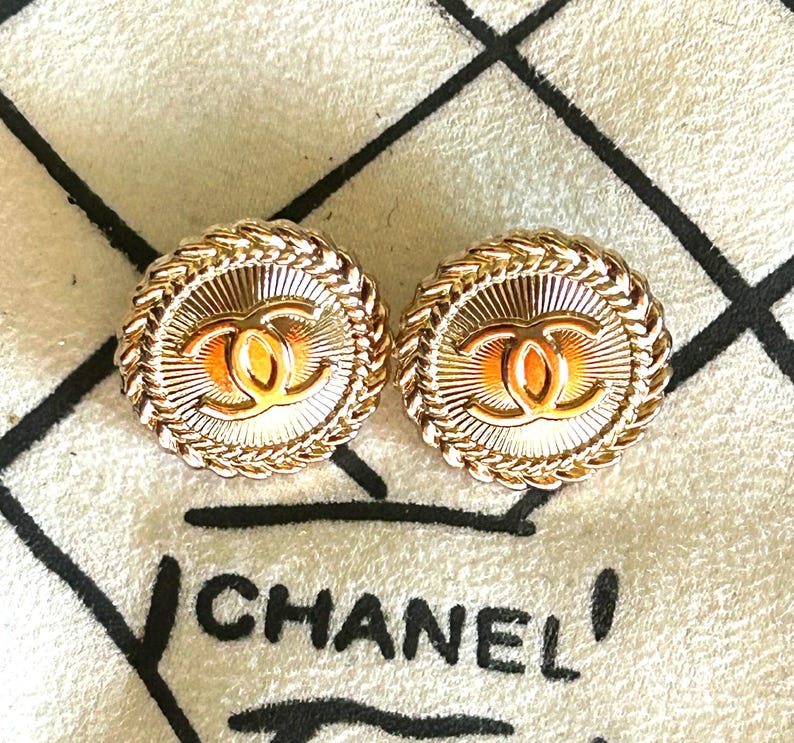 May include: A pair of round gold-tone earrings featuring a central interlocking CC logo surrounded by a sunburst pattern and a braided rope border. The earrings are displayed against a textured off-white fabric with the black printed text CHANEL.