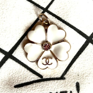 May include: A white flower-shaped pendant with gold trim and a pink gemstone center. The pendant features a double-C logo at the base of the flower. The pendant is on a white surface with a black and white pattern.