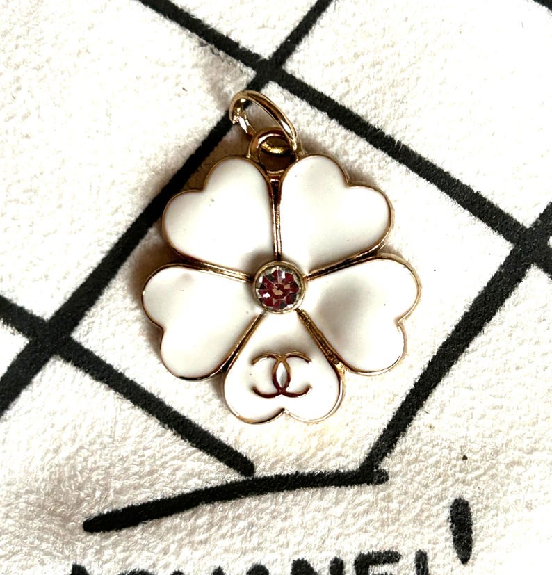 May include: A white flower-shaped pendant with gold trim and a pink gemstone center. The pendant features a double-C logo at the base of the flower. The pendant is on a white surface with a black and white pattern.