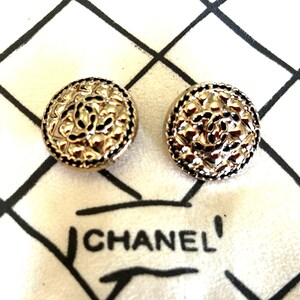 May include: Two gold-toned, round earrings with a floral design and black accents. The earrings are on a white fabric with a black grid pattern and the word "CHANEL" printed on it.