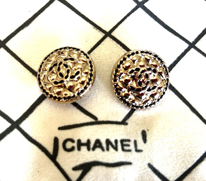 May include: Two gold-toned, round earrings with a floral design and black accents. The earrings are on a white fabric with a black grid pattern and the word "CHANEL" printed on it.