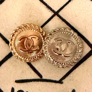 May include: Two circular metal buttons featuring the interlocking CC logo in the center. Each button has a sunburst pattern radiating from the logo and a braided rope-style border. One button is gold-toned, and the other is silver-toned.