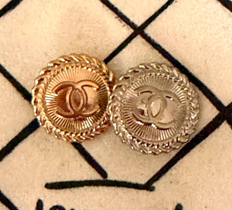 May include: Two circular metal buttons featuring the interlocking CC logo in the center. Each button has a sunburst pattern radiating from the logo and a braided rope-style border. One button is gold-toned, and the other is silver-toned.
