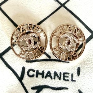 May include: A pair of circular gold-tone earrings featuring the text CHANEL PARIS in a cut-out design. The center of each earring displays the iconic interlocking CC logo encrusted with clear crystals, creating a reflective and metallic appearance.