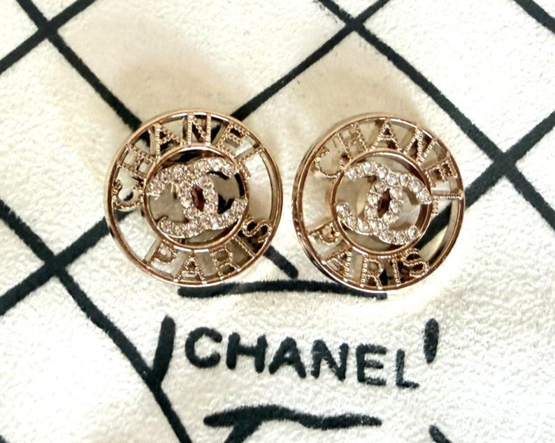 May include: A pair of circular gold-tone earrings featuring the text CHANEL PARIS in a cut-out design. The center of each earring displays the iconic interlocking CC logo encrusted with clear crystals, creating a reflective and metallic appearance.