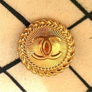 May include: A round gold-tone metal button featuring a central embossed interlocking CC logo. The design includes a sunburst pattern radiating from the center and a braided rope-style border around the outer edge of the circular button.