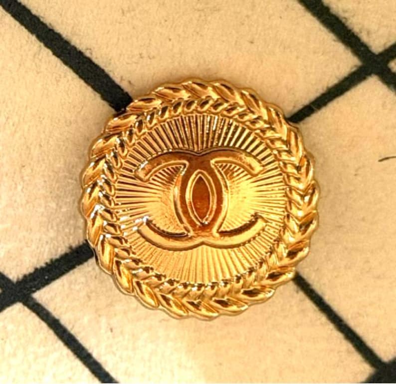 May include: A round gold-tone metal button featuring a central embossed interlocking CC logo. The design includes a sunburst pattern radiating from the center and a braided rope-style border around the outer edge of the circular button.