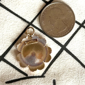 May include: A gold-toned flower-shaped charm with the word "CHANEL" on it. The charm has a textured surface and a small loop at the top. A US quarter is next to the charm.