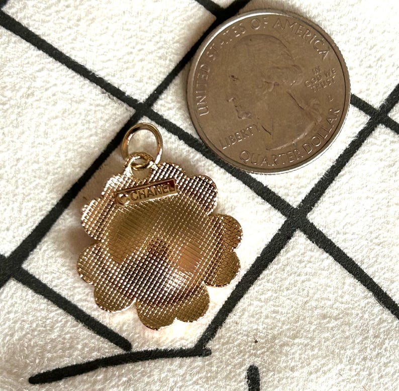 May include: A gold-toned flower-shaped charm with the word "CHANEL" on it. The charm has a textured surface and a small loop at the top. A US quarter is next to the charm.