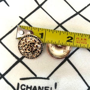 May include: Two gold-toned buttons with intricate floral designs. The buttons are approximately 1 inch in diameter. A yellow measuring tape is placed over the buttons, showing the measurement in inches.