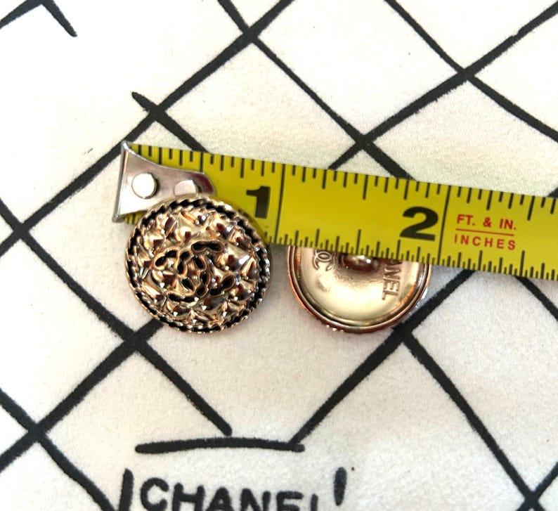May include: Two gold-toned buttons with intricate floral designs. The buttons are approximately 1 inch in diameter. A yellow measuring tape is placed over the buttons, showing the measurement in inches.
