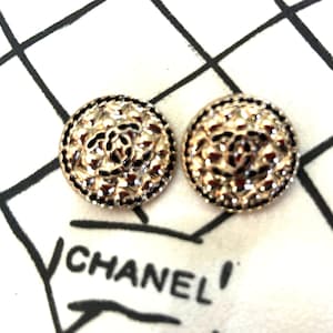 May include: Two gold-toned, round earrings with a textured surface and a black border. The earrings feature a central design and are displayed on a white surface with the word "CHANEL" printed in black.