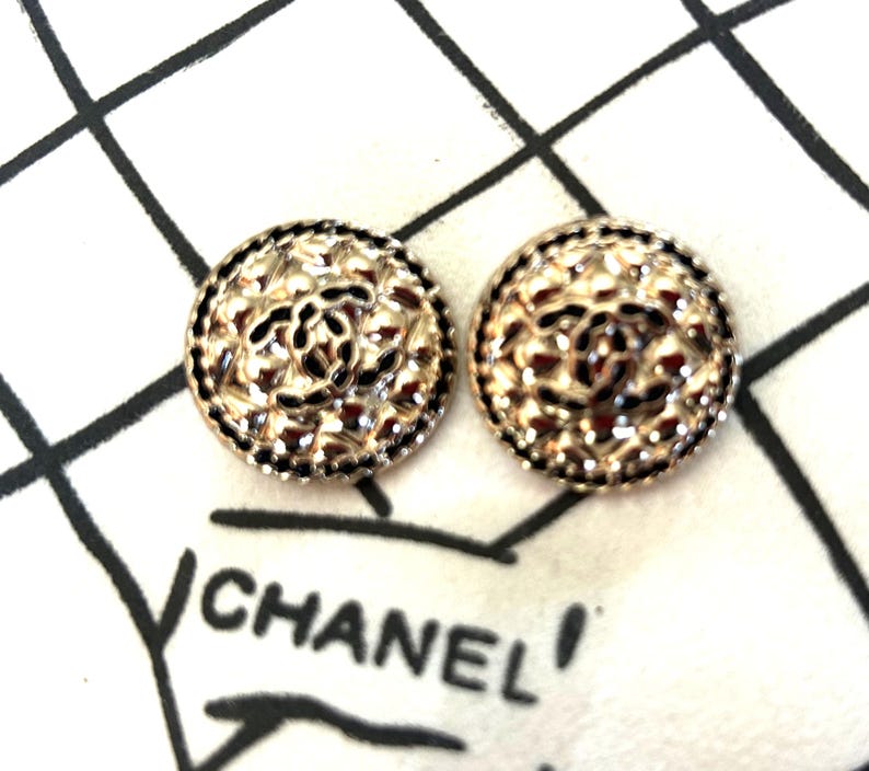May include: Two gold-toned, round earrings with a textured surface and a black border. The earrings feature a central design and are displayed on a white surface with the word "CHANEL" printed in black.