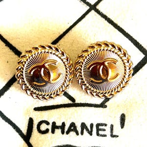 May include: A pair of round gold-tone metal earrings featuring a central interlocking CC logo, a sunburst textured background, and a braided rope-style border. The earrings are displayed against a white background with the black text CHANEL.