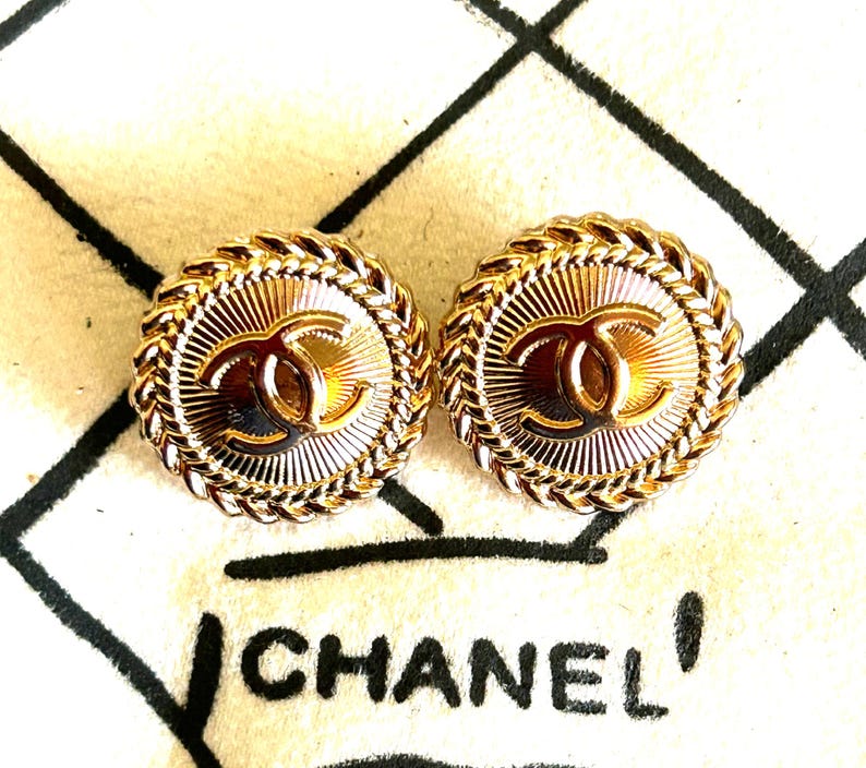 May include: A pair of round gold-tone metal earrings featuring a central interlocking CC logo, a sunburst textured background, and a braided rope-style border. The earrings are displayed against a white background with the black text CHANEL.