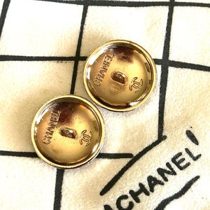 May include: Two gold-toned CHANEL buttons with a round shape. Each button has the brand name and logo embossed on the back. The buttons are on a white fabric with a black CHANEL logo.