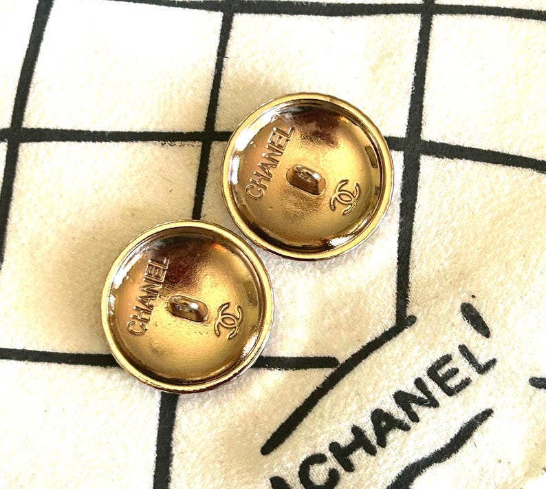 May include: Two gold-toned CHANEL buttons with a round shape. Each button has the brand name and logo embossed on the back. The buttons are on a white fabric with a black CHANEL logo.