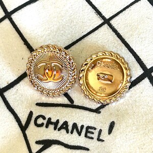 May include: Two round gold-tone buttons featuring a central interlocking CC logo and a sunburst pattern, surrounded by a braided rope border. The reverse side of one button displays the text CHANEL and a small CC logo with a metal shank attachment.
