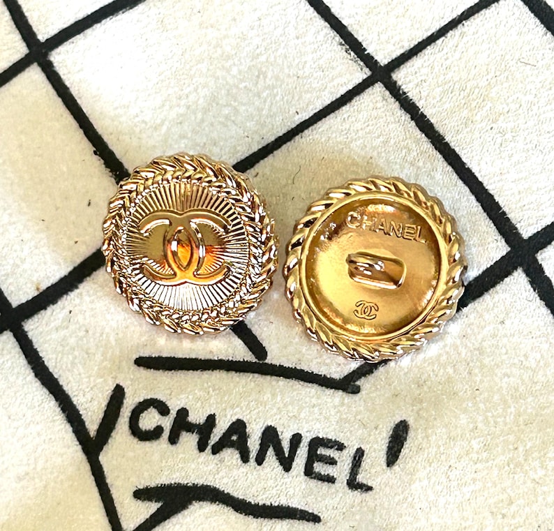 May include: Two round gold-tone buttons featuring a central interlocking CC logo and a sunburst pattern, surrounded by a braided rope border. The reverse side of one button displays the text CHANEL and a small CC logo with a metal shank attachment.