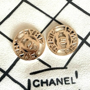 May include: Two circular gold-toned buttons featuring a cut-out design with the interlocking CC logo in the center. The outer rim of each button displays the text CHANEL in a repeating pattern. The buttons have a smooth, metallic finish.