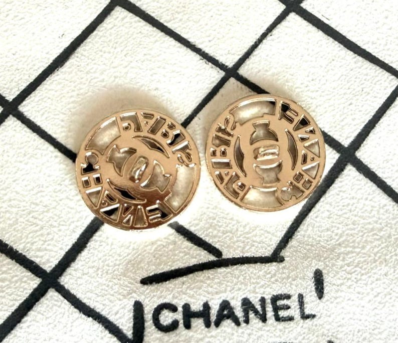 May include: Two circular gold-toned buttons featuring a cut-out design with the interlocking CC logo in the center. The outer rim of each button displays the text CHANEL in a repeating pattern. The buttons have a smooth, metallic finish.