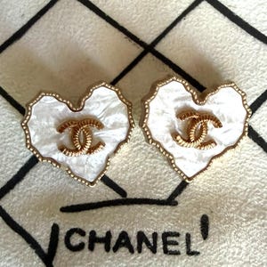 May include: A pair of heart-shaped earrings featuring a white pearlescent finish with a gold-toned scalloped edge. Each earring displays a central gold-toned interlocking CC logo. The design is consistent with the Chanel brand aesthetic.