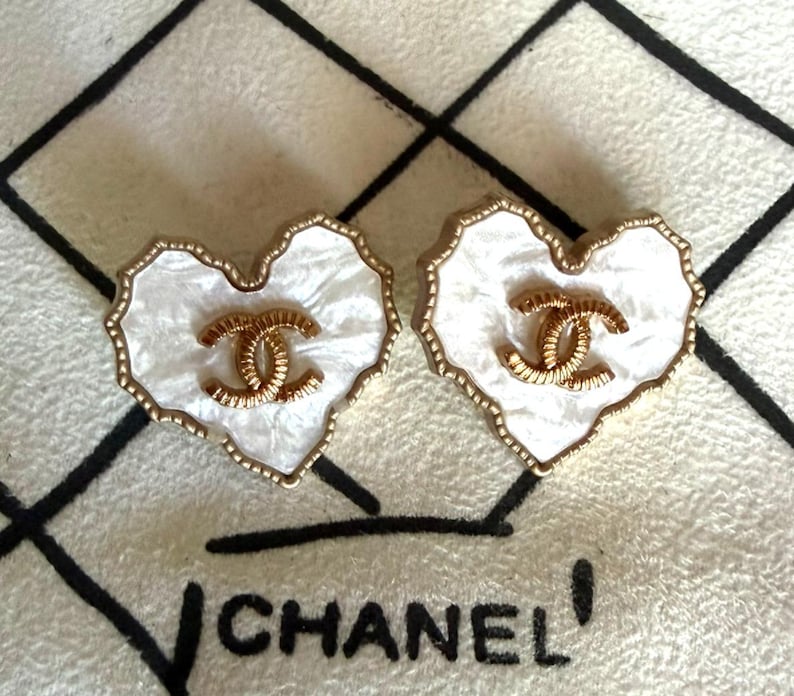 May include: A pair of heart-shaped earrings featuring a white pearlescent finish with a gold-toned scalloped edge. Each earring displays a central gold-toned interlocking CC logo. The design is consistent with the Chanel brand aesthetic.
