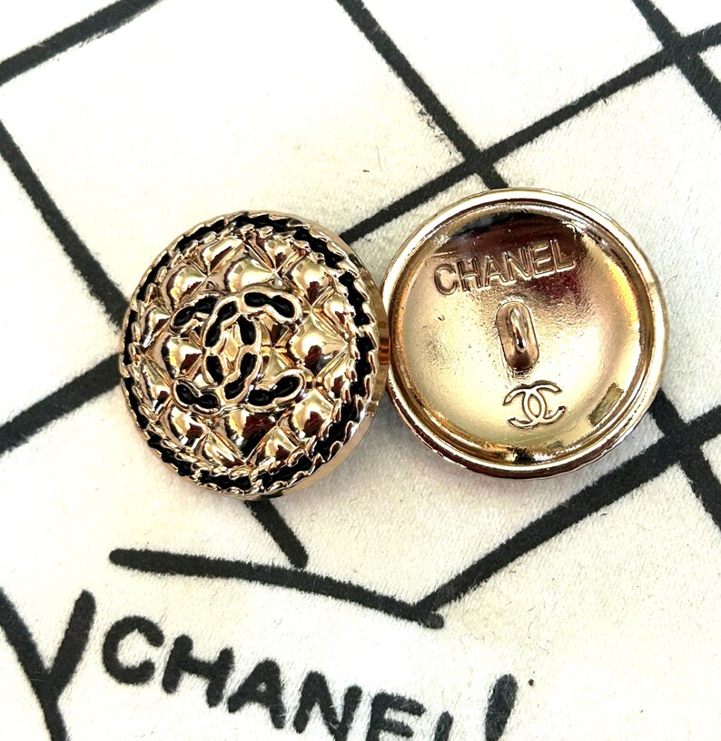 May include: Two gold-toned buttons with the brand name "CHANEL" visible. One button features a textured, ornate design with black accents. The other button displays the brand name and logo on the back. The buttons are round.