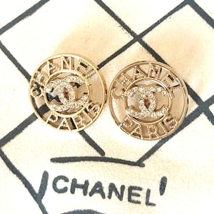 May include: A pair of circular gold-tone earrings featuring the text CHANEL PARIS in a cut-out design surrounding a central interlocking CC logo embellished with clear crystals. The earrings are displayed against a cream fabric background.