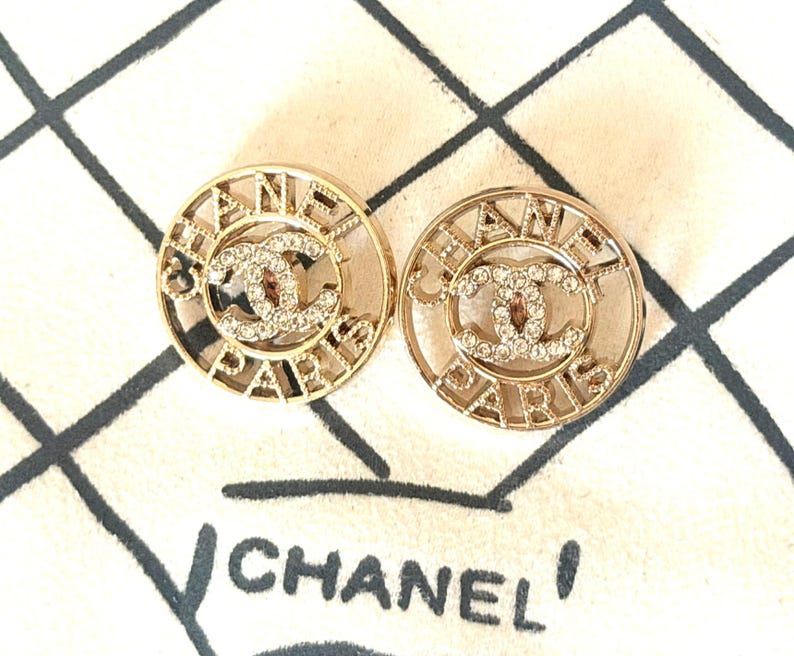 May include: A pair of circular gold-tone earrings featuring the text CHANEL PARIS in a cut-out design surrounding a central interlocking CC logo embellished with clear crystals. The earrings are displayed against a cream fabric background.