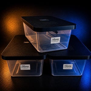 May include: Three clear plastic storage containers with black lids are stacked. Each container has a rectangular shape and a label that reads "Slavens". The lids have a textured surface. The background has orange and blue lighting.