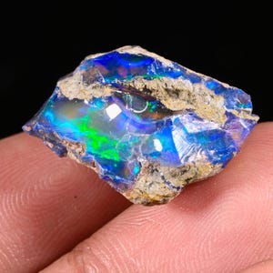 May include: A raw, uncut opal gemstone displaying vibrant flashes of electric blue and neon green play-of-colour. The stone features a rough, textured, earthy brown matrix running through the centre, contrasting with the polished, translucent, and reflective crystalline sections of the mineral specimen.