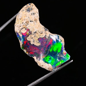May include: A raw, irregular boulder opal specimen featuring a rough, sandy-colored matrix exterior and a polished, vibrant interior face displaying intense flashes of neon green, electric blue, and deep red play-of-color against a dark base.