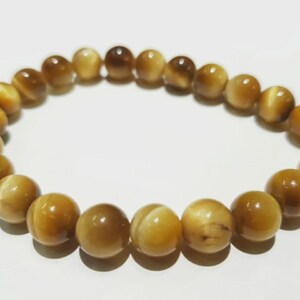 Pode incluir: Uma pulseira de contas de olho de tigre dourado. A pulseira apresenta contas redondas e polidas em tons de ouro e castanho. As contas são enfiadas para formar uma forma circular, perfeita para o pulso.