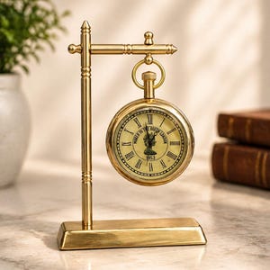 May include: A gold-toned metal desk clock suspended from a matching stand. The round clock face features Roman numerals, a central silhouette profile, and the text "MAKER TO THE QUEEN LONDON 1953". The stand has a rectangular base and a vertical post.