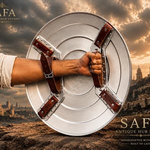 May include: A circular silver-toned metal shield featuring a smooth, concentric ring design. The back of the shield is equipped with two brown leather straps secured by metal rivets and a buckle, designed for forearm and hand attachment.