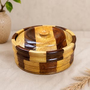 May include: A round wooden container with a lid, featuring a contrasting checkerboard pattern of light tan and dark brown wood segments. The container has a smooth, polished finish and a small, centered circular knob on the top of the lid.