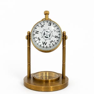 May include: A vintage-style brass compass on a stand. The compass has a white face with black directional markings and a needle. The base and supporting structure are also brass. The compass is designed for decorative purposes.