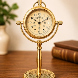 May include: A gold-toned metal desk clock with a circular face featuring both 12-hour and 24-hour time markings. The clock is mounted on a vertical stand with a circular base decorated with an intricate embossed floral pattern and a top crown.