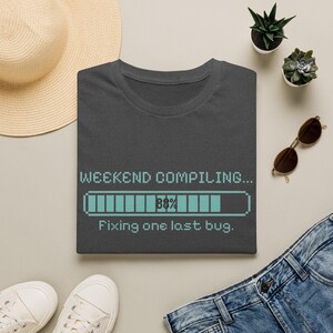 Può includere: Una t-shirt grigio scuro con una grafica a barra di caricamento pixelata azzurra con il testo "WEEKEND COMPILING..." sopra e "88%" all'interno della barra, seguito dal testo "Fixing one last bug." sotto la barra di caricamento.