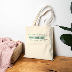 May include: A cream-colored canvas tote bag featuring a green pixelated progress bar graphic. The text reads "WEEKEND COMPILING..." above the bar, "88%" inside the bar, and "Fixing one last bug." below the bar. The bag has two long shoulder straps.