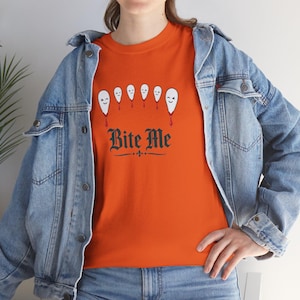 Puede incluir: Una camiseta naranja con un gráfico de seis figuras blancas similares a fantasmas con gotas rojas que parecen sangre, situadas sobre el texto negro "Bite Me" en fuente gótica, usada debajo de una chaqueta de mezclilla azul claro.