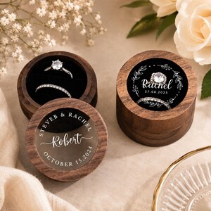 May include: Two round wooden ring boxes with dark wood grain and clear lids. One lid features the text "STEVEB & RACHEL", "Robert", and "OCTOBER 15, 2024". The other box contains a diamond ring and a band, with a black insert showing "Rachel" and "27.08.2025".