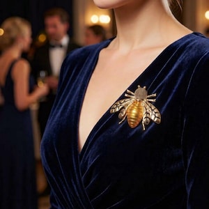 May include: A gold-toned bee brooch featuring a textured head and a smooth, rounded abdomen, pinned to a deep navy blue velvet dress with a V-neckline. The insect-shaped accessory displays detailed wing patterns and a metallic finish.