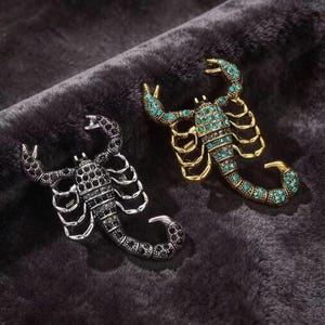 May include: Two scorpion-shaped brooches. One brooch is silver-toned with black crystals covering its body and pincers. The second brooch is gold-toned with teal crystals embedded across its segmented body, legs, and curved tail structure.