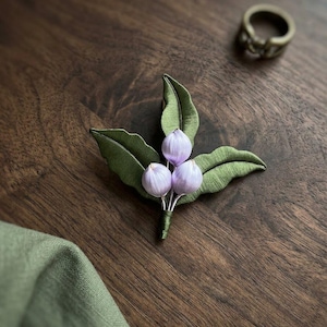 Può includere: Una spilla in tessuto con tre piccoli boccioli arrotondati color lavanda pallido raggruppati con quattro foglie verdi testurizzate. I gambi sono avvolti in filo verde e un anello metallico scuro è posizionato nelle vicinanze.